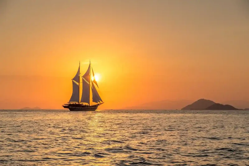 A phinisi in silhouette against an orange sunset on open water
