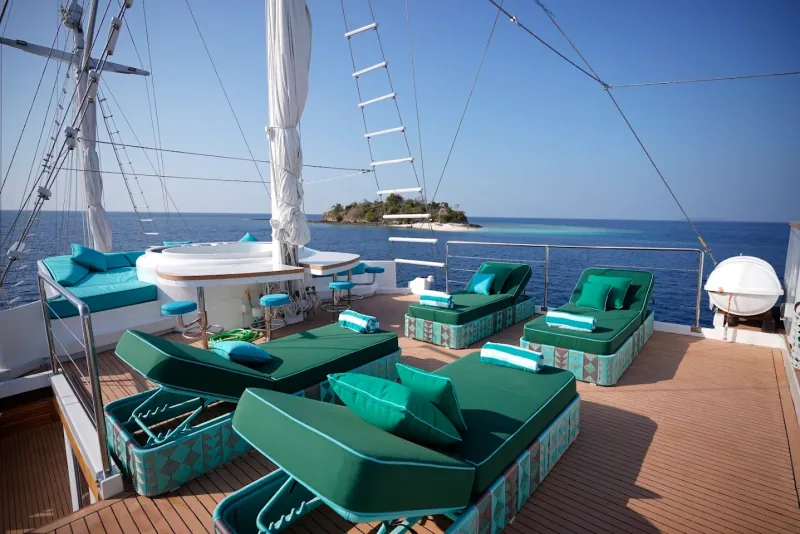 The sundeck of a phinisi with green loungers on a teak deck, an island on the horizon
