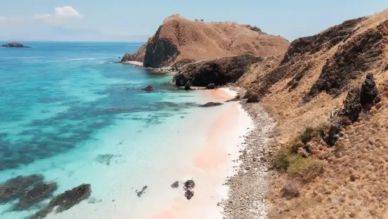 Aerial view of Pink Beach: pale pink sand curving between turquoise shallows and dry brown hills